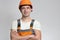 Confident young construction worker in hard hat and overalls with arms folded on the chest on grey studio background