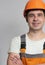 Confident young construction worker in hard hat and overalls with arms folded on chest on grey studio background