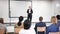 Confident young female lecturer standing with microphone on stage in conference room, speaking to businesspeople at