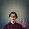 Confused adolescent guy, wearing eyeglasses gesturing, pointing index finger up, perplexed expression isolated on grey wall