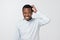 Confused african man scratching head while looking at camera and standing over gray background.