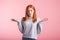 Confused redhead girl shrugs shoulders in studio on pink background.