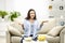 Confused, but smiling brunette woman doesn`t know which snack to choose, isolated, sitting on the couch at home.