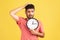 Confused thoughtful man with beard in red striped t-shirt rubbing his head holding in hands big wallclock, time out, worried about