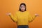 Confused uncertain asian woman shruging unsurely while wearing yellow sweater on orange background.