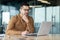 Confused young male businessman sits in the office in front of a laptop and looks thoughtfully at the screen