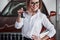 Congratulations with purchase. Woman in eyewear and white shirt stands near the red automobile