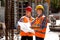 Construction manager and engineer dressed in orange work vests and hard helmets explore construction documentation on