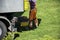 Construction worker in orange overall stands behind a truck on a grass field and fills a wheelbarrow