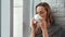 Contemplative domestic young woman drinking coffee looking through window enjoying loneliness