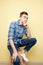 Contemplative guy dressed in a plaid shirt and jeans is sitting on a stool on a yellow background in the studio