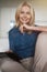 Content mature woman smiling at camera, using digital tablet pc while relaxing on a couch at home in the living room