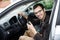 Contented young driver is sitting at his car while looking at the camera. He is holding the keys at his left hand. His right hand