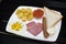 Continental breakfast â€“ one egg omlet, baked potato; backed tomato; two pics bread, sausage and meyones on black wood table. Top