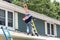 Contractor carrying a package of roof shingles on a roof with ladders