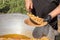 Cook puts pilaf with meat and carrots on a disposable paper plate against the backdrop of a large cast iron cauldron.