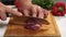 A cook is slicing beef meat on a kitchen board. In the background is a red bell bell pepper and a bunch of parsley