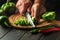 The cook slicing fresh green peppers on a wooden cutting board. Close-up of chef hands while preparing vegetarian food or adjika