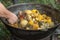 cook uses a slotted spoon to stir meat, corn on the cob, potatoes and vegetables in a cast-iron cauldron on an outdoor wood-