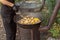 cook uses a slotted spoon to stir meat, corn on the cob, potatoes and vegetables in a cast-iron cauldron on an outdoor wood-