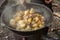 cook uses a slotted spoon to stir meat, corn on the cob, potatoes and vegetables in a cast-iron cauldron on an outdoor wood-