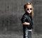 Cool brutal kid girl in leather jacket, jeans and sunglasses is posing like a biker rockstar on concrete wall background