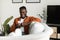 Cool gadget and app. Excited black man holding mobile phone and typing sms message, resting on sofa at home, free space