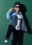 Cool kid boy in blue sunglasses, headwear, fleece jacket, pants is acting like a hip-hop star, showing cool sing on green