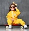 Cool kid girl in yellow hooded jumpsuit costume, sunglasses and killing yellow bow sits on cool red skateboard with playful smile