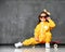 Cool kid girl in yellow hooded jumpsuit costume, sunglasses and yellow bow sits in cool pose on red skateboard