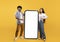 Cool mobile offer. Happy indian couple pointing at big cellphone with white screen for mockup, yellow background