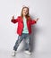 Cool and positive blond baby girl kid in red leather jacket, white t-shirt and blue jeans gestures V-signs victory