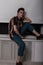 Cool stylish young man in a striped vintage shirt in blue youth jeans is resting near the window in the room in the evening.