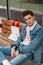 Cool stylish young man in trendy blue youth casual jeans clothes sitting on wooden bench and waiting for public transport.