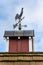 Copper rooster weathervane on top of red rooftop cupola with a blue sky and white clouds in the background