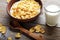 Cornflakes in a brown clay plate on a worn wooden background, next is a glass of milk and a spoon