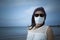 Coronavirus seaside holidays: half-length shot of a woman at the beach look at the camera with the mask for Covid-19 pandemic with