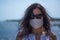 Coronavirus seaside holidays: half-length shot of a woman at the beach look at the camera with the mask for Covid-19 pandemic with
