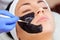 Cosmetologist does a black mask to cleanse the skin and anti-wrinkle on the face of a beautiful woman, spa treatments and