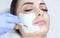 The cosmetologist for the procedure of cleansing and moisturizing the skin, applying a mask with stick to the face of a young woma