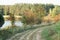 Country road along weeds, lake, pine forest and wild growing trees with red berries of haw thorn