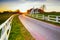 Country road in dutch countryside with white fence leading towards an old farmhouse in Holland during an atmospheric sunset