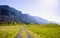 Country road overgrown with green grass leads to a mountainous rocky ridge along the river through a meadow and a strip of bright