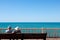 A couple of adults sits on the promenade and looks out at the sea