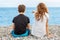 Couple of beautiful teens, first love. Guy hugs a girl sitting on the pebble beach next to each other and looking at the sea.