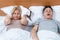 Couple in bed, man sleeping and snoring with open mouth, disgruntled woman covering her ears with pillow. Snoring as a problem.