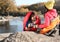 Couple of campers in sleeping bags sitting on rock near pond.