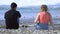 Couple drinking wine on stone beach. Media. Young man and woman drink wine sitting on rocky shore near blue sea. Young