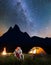 Couple hikers sitting together near campfire and glowing tent at night under stars and looking to the starry sky