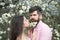 Couple in love at blooming garden. Tender woman with long dark hair and handsome man with tiny white flowers in beard